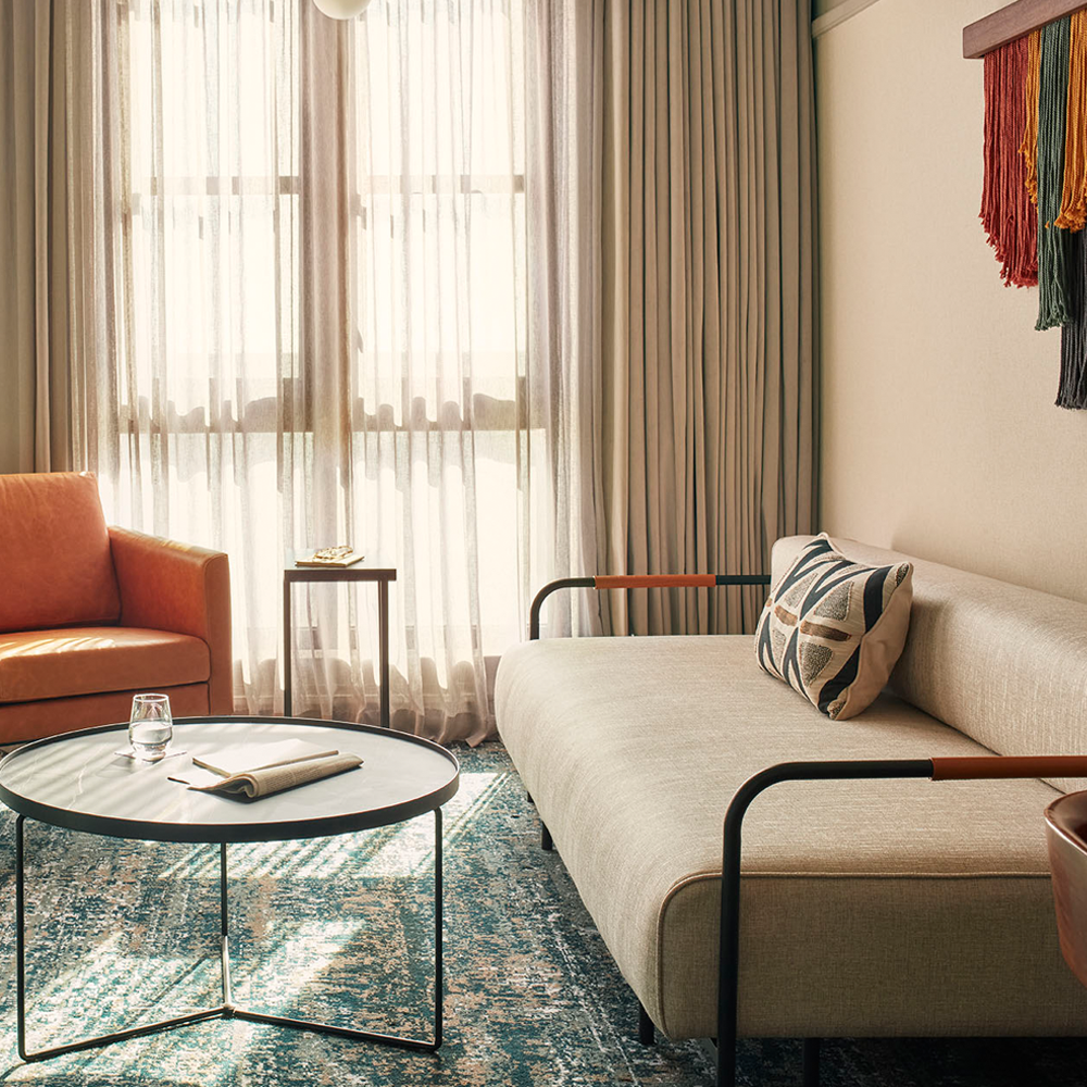 Sunlight pouring through the sheer curtains, a 1-seater, leather brown couch with a side table, and a 3-seater couch to the right of the image. There is a black, round table in front of the couches.