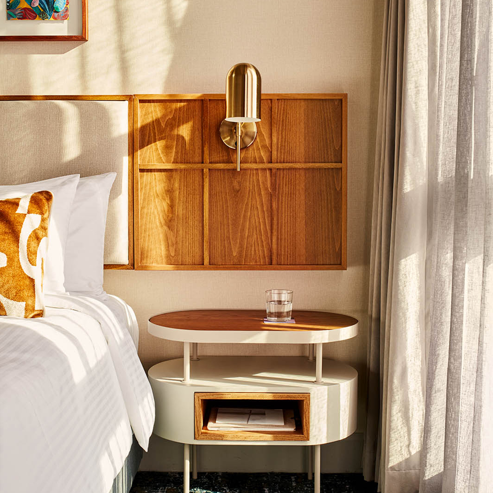 A well-lit, close-up image of the bedside pedestal, with art pieces hanging on top of the bed, and the cream headboard.