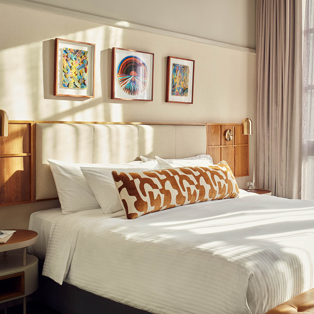 A well-lit image of the king-sized bed, with art pieces hanging on top of the bed, and the cream headboard.