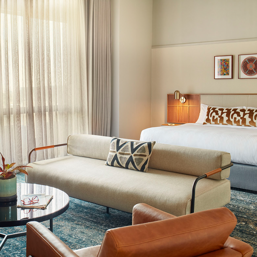 A lounging area, with a 1-seater brown, leather couch, and a 3-seater couch surrounding the black, round table. There is a king-sized bed in the back, with art pieces on top of the bed.