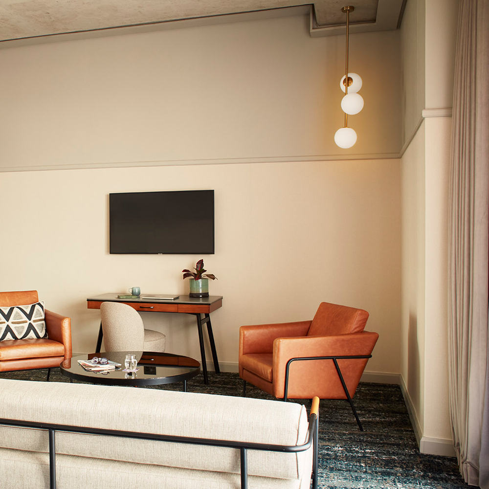 A TV hanging above the desk, which can be used for work or as a makeup area, along with two 1-seater brown, leather couches and is surrounding the black, round table.