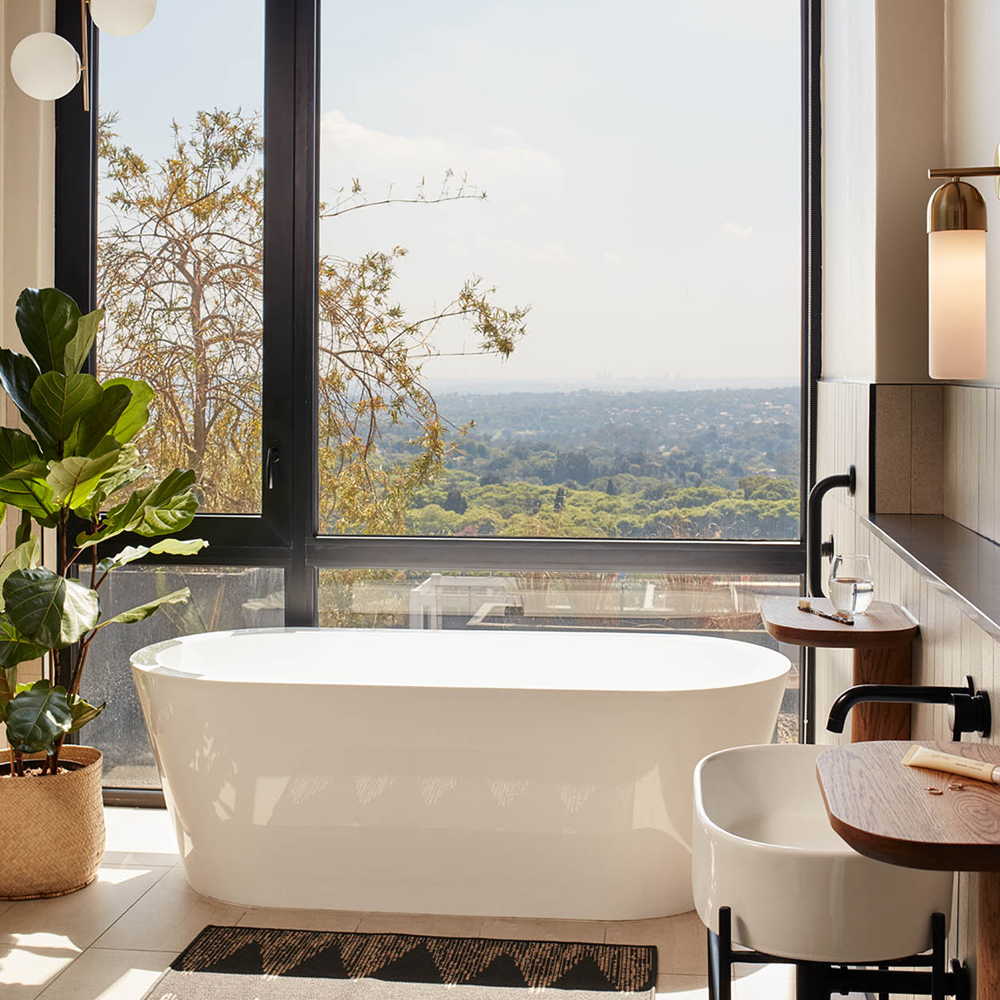 Floor to ceiling windows, with a bathtub in front, and a white sink, with lights on either side of the mirror.