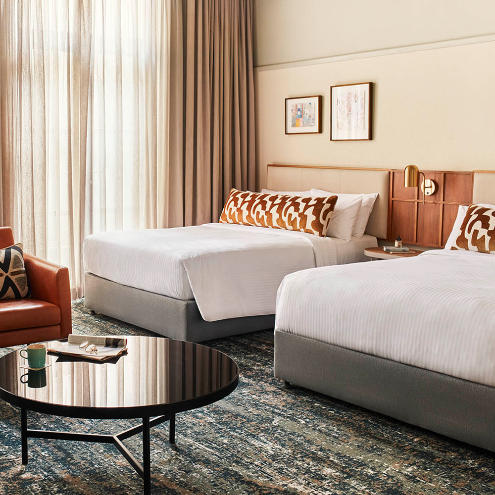 Two double-sized beds, with a 1-seater brown, leather couch. A black round table in front of the beds, and sunlight pouring through the sheer curtains in the back.