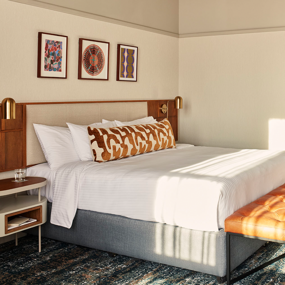 A well-lit image of the king-sized bed, with art pieces hanging on top of the bed, and the cream headboard.