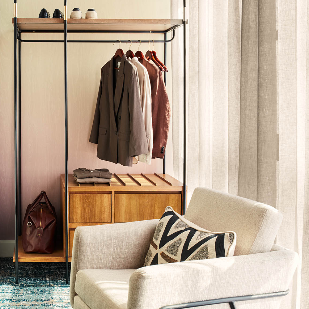 Sunlight pouring through, onto the 1-seater grey couch. Behind the couch, there is an open wardrobe with wooden drawers at the bottom.