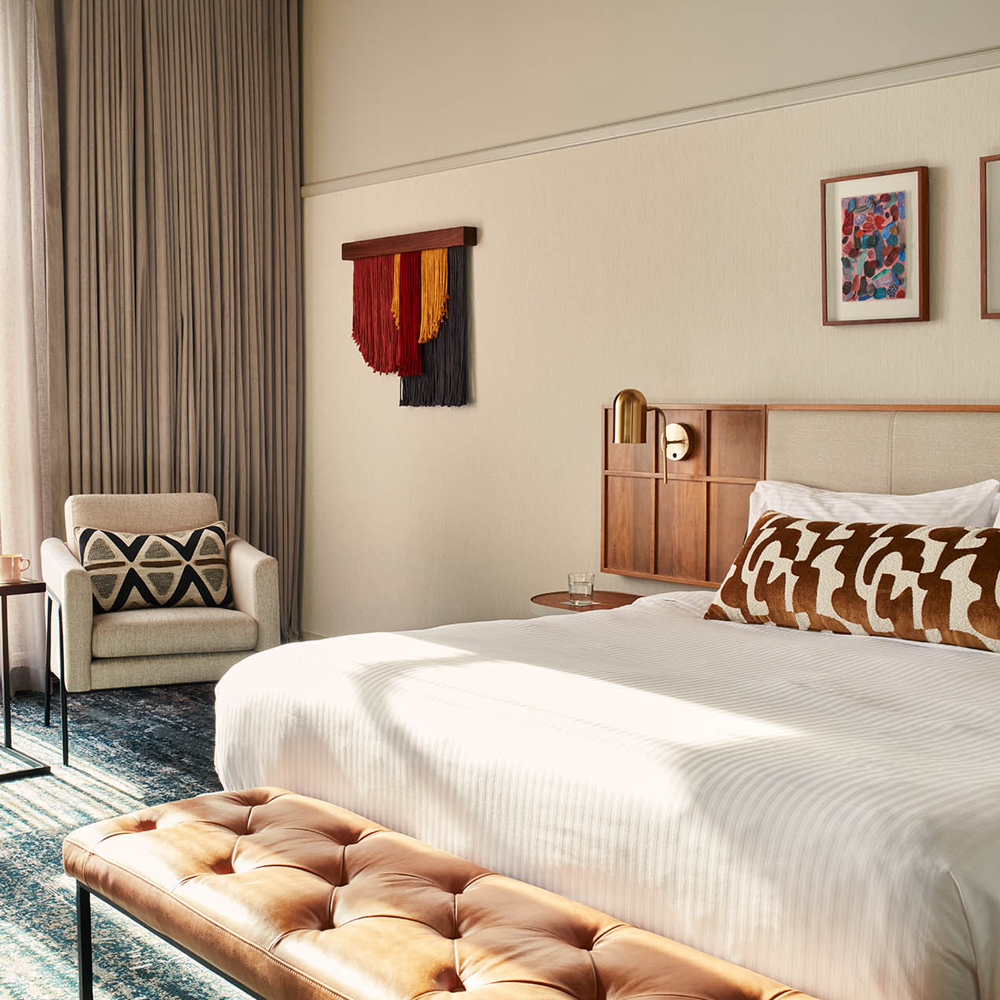 Sunlight pouring through the room, a 1-seater couch on the left of the image, and a king-sized bed on the right, with some art hanging on the wall.