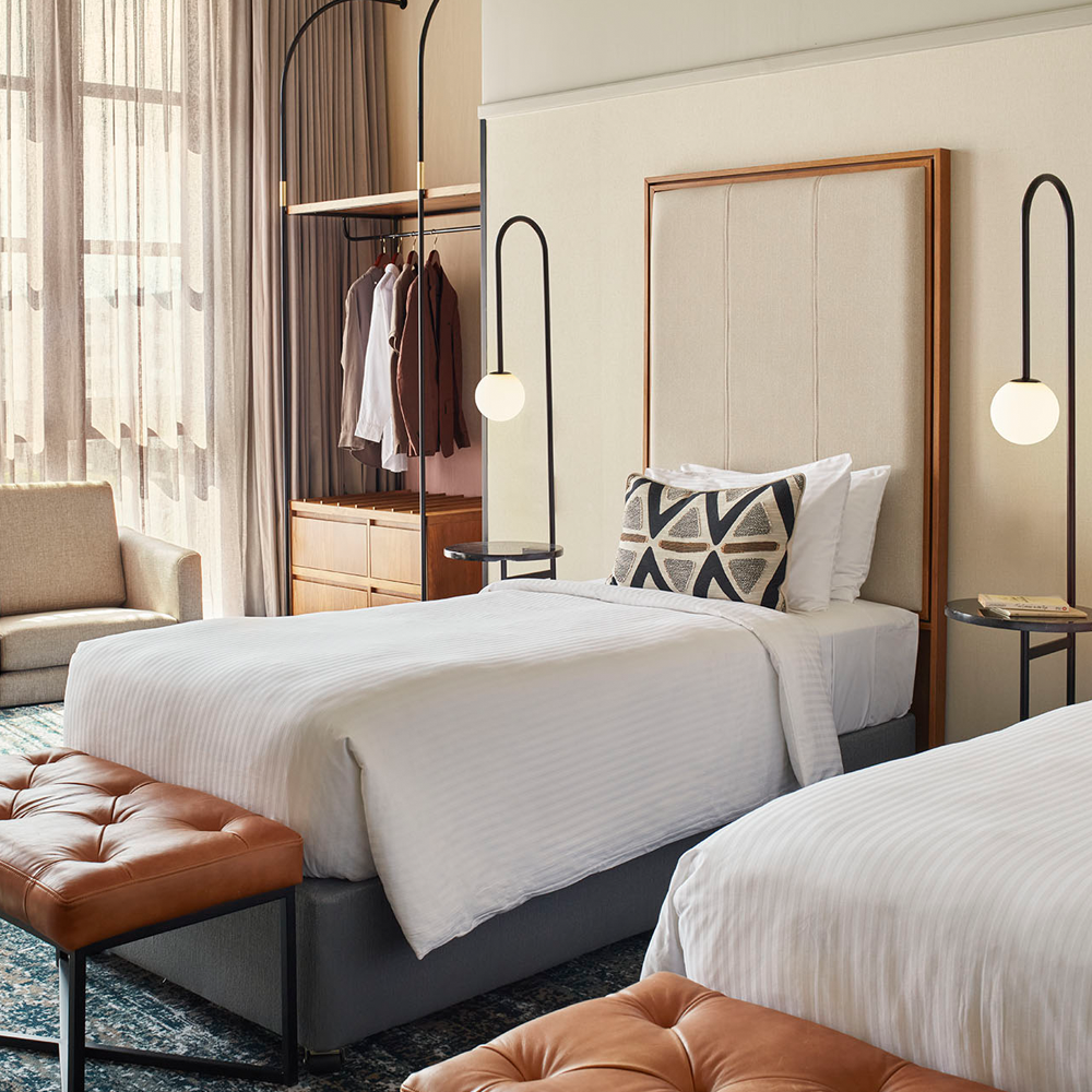 Two twin-sized beds, each with a light on the side of the bed, and sunlight pouring through the sheer curtains on to the 1-seater grey couch, and the open wardrobe with wooden drawers on the bottom.