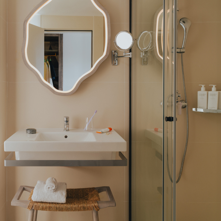 A bathroom with large mirror and smaller vanity mirror, sink and chair, with shower