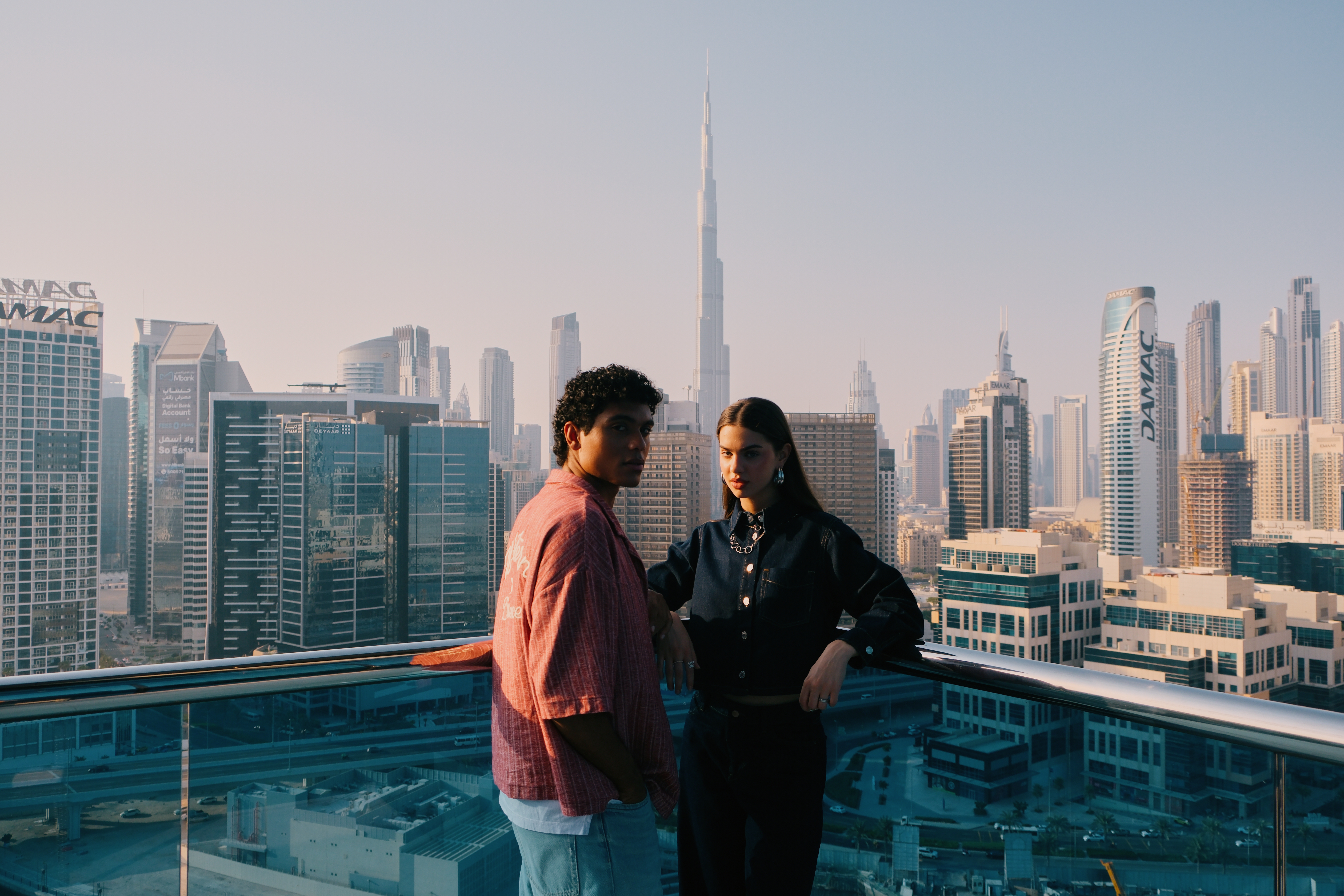 A man and a woman standing on a balcony with the Dubai city skyline, including the Burj Khalifa and other skyscrapers, in the background.