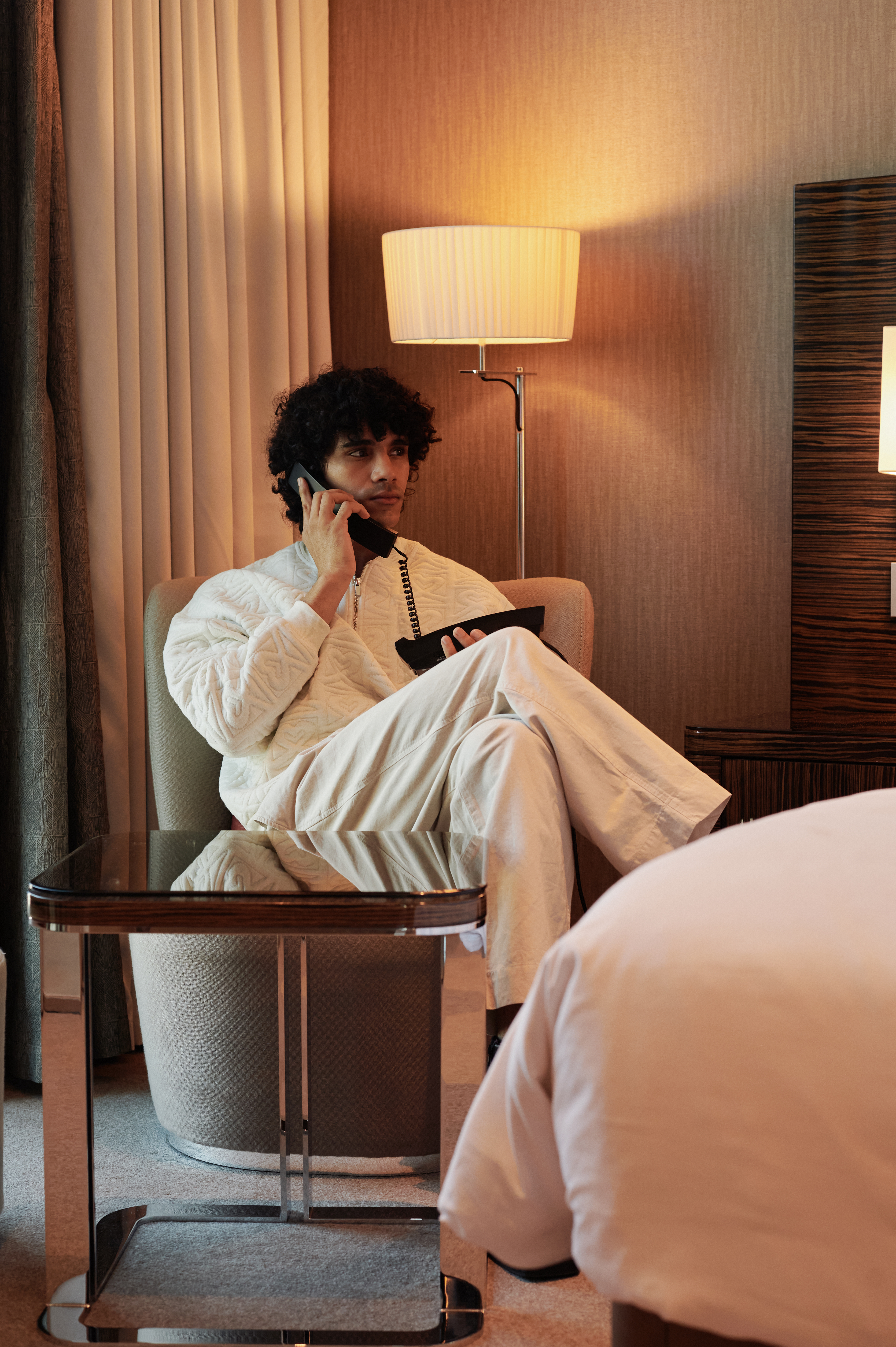 A man sitting in an armchair in a room, holding a landline phone and speaking