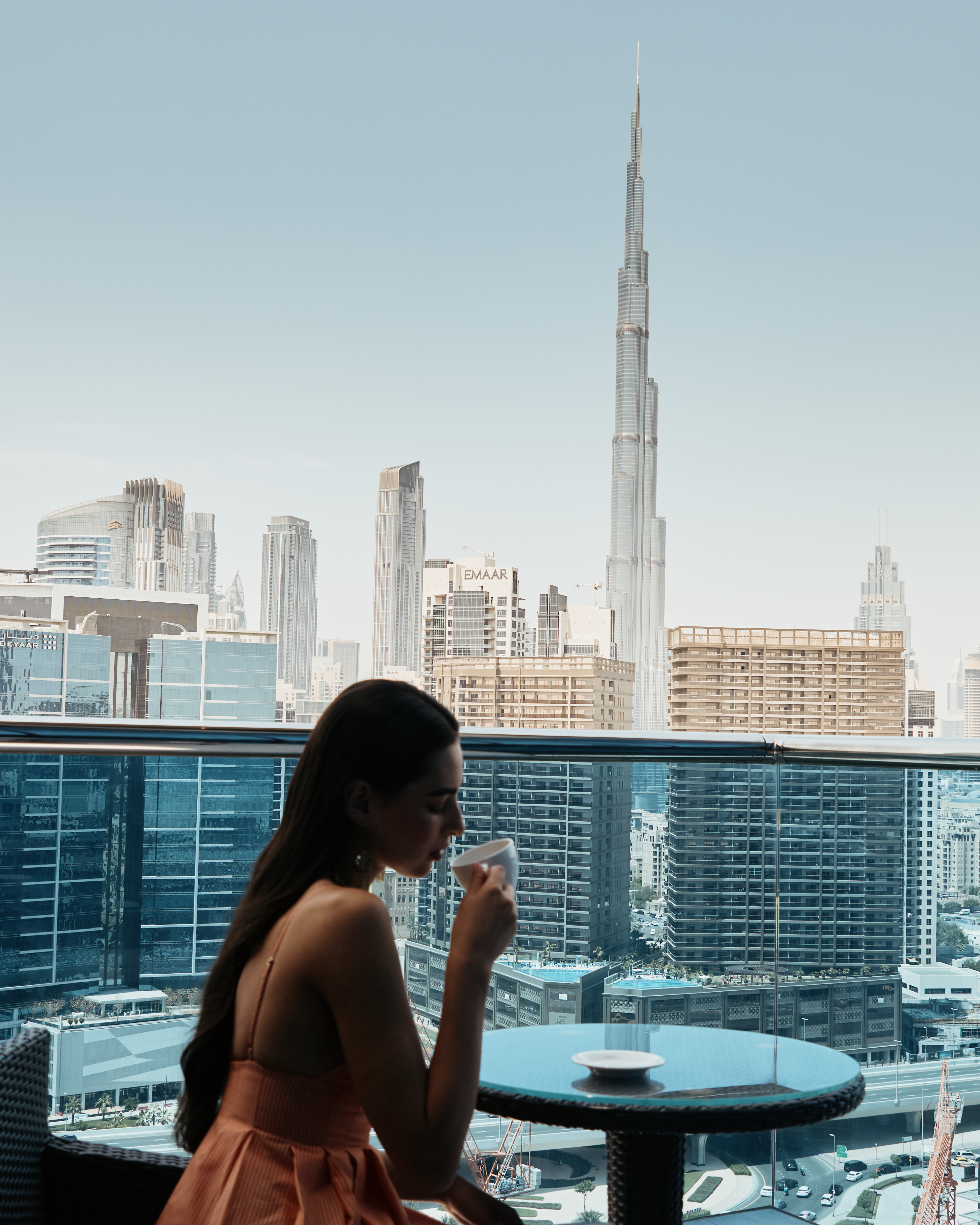 A woman sitting on the balcony of a hotel room with a stunning view of the Burj Khalifa, holding a cup and enjoying a drink