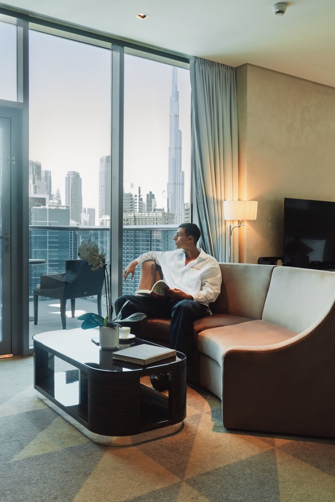 A man sitting on a couch in a hotel room, by the window and balcony, with scenic views of the Dubai skyline, including the Burj Khalifa and other towering skyscrapers
