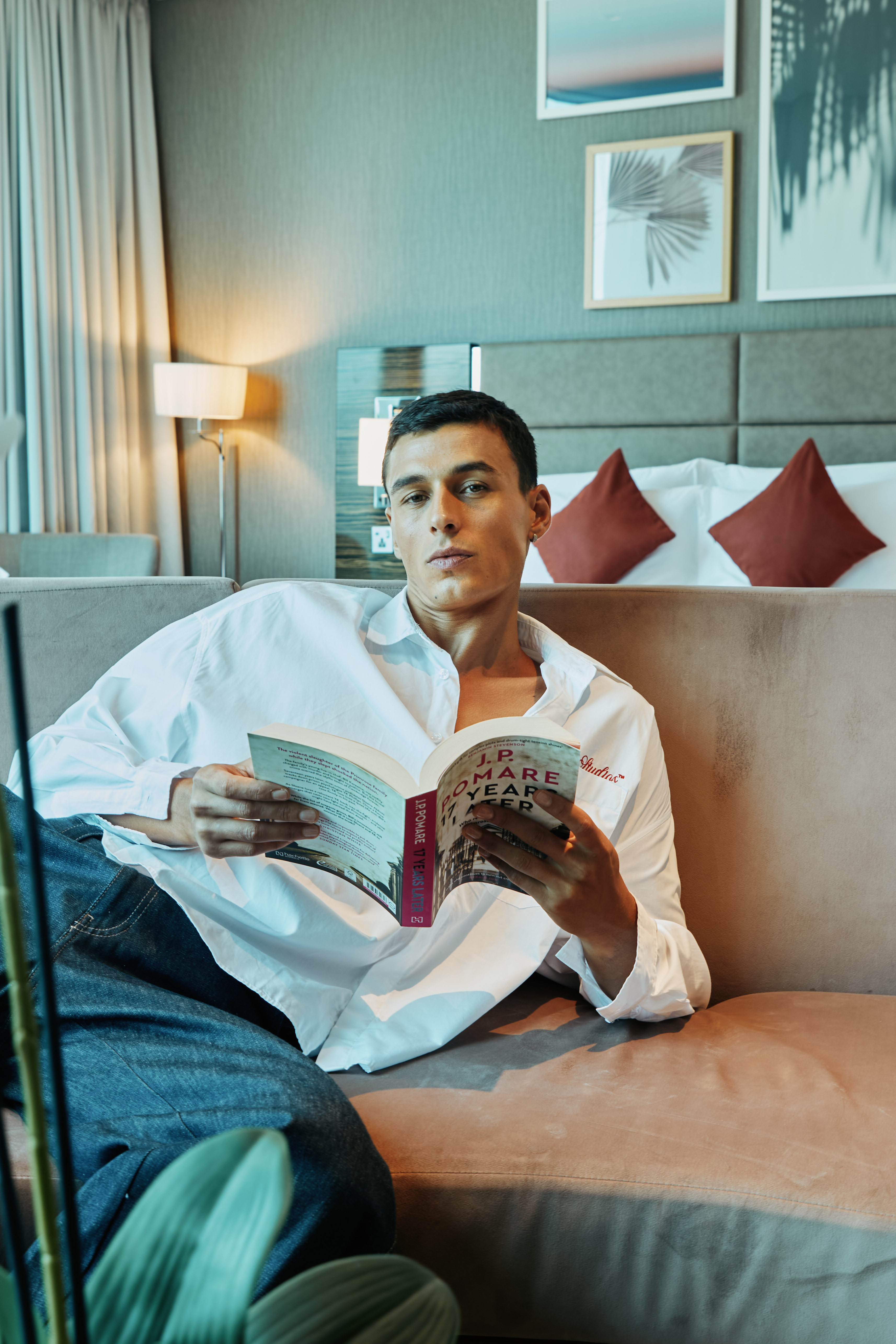 A man sitting on a couch in a hotel room, holding a book and looking relaxed.