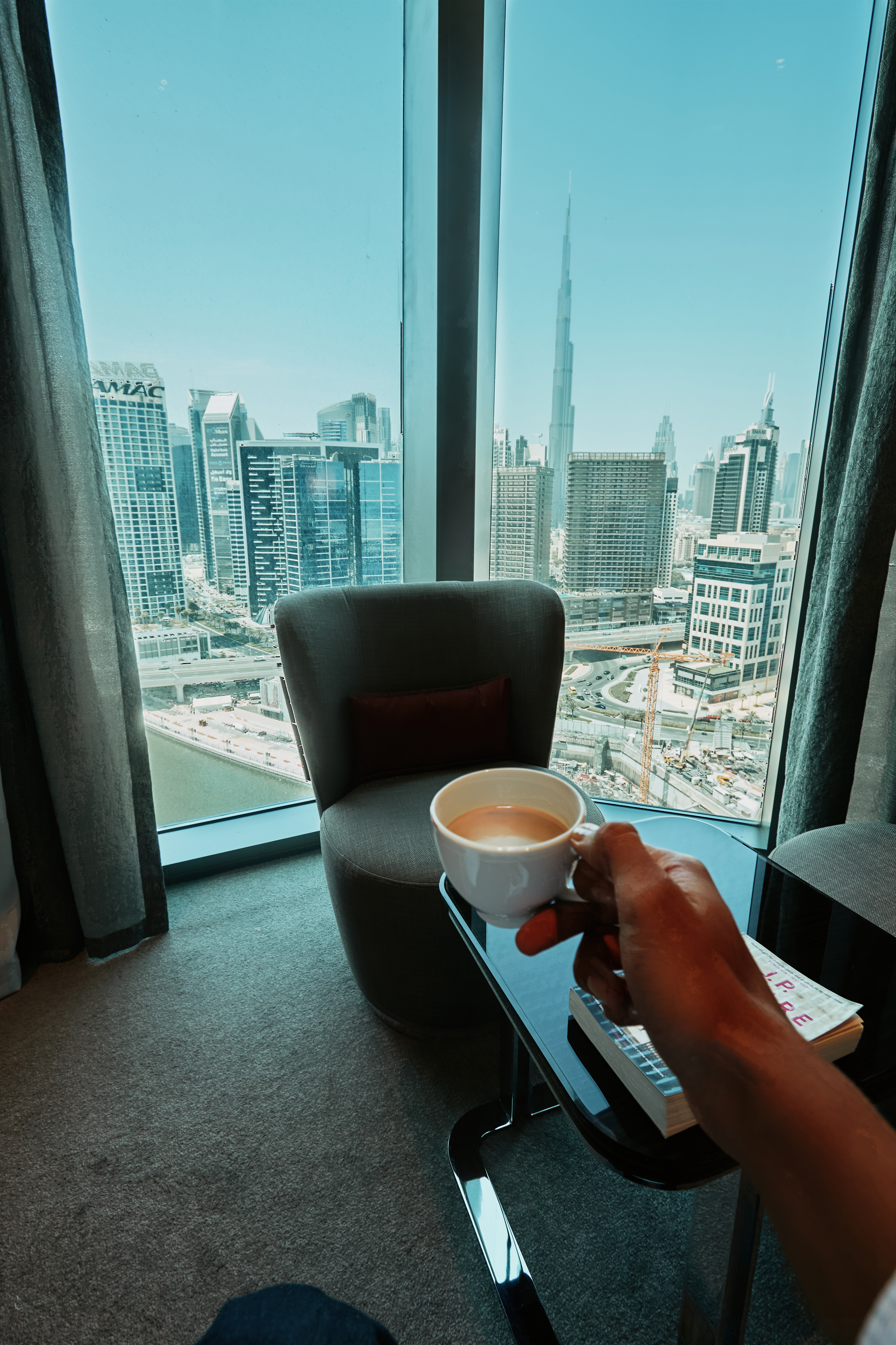A guest in a hotel room holding a cup of coffee, standing next to a large window with stunning views of the Dubai skyline, including the Burj Khalifa and other towering skyscrapers.