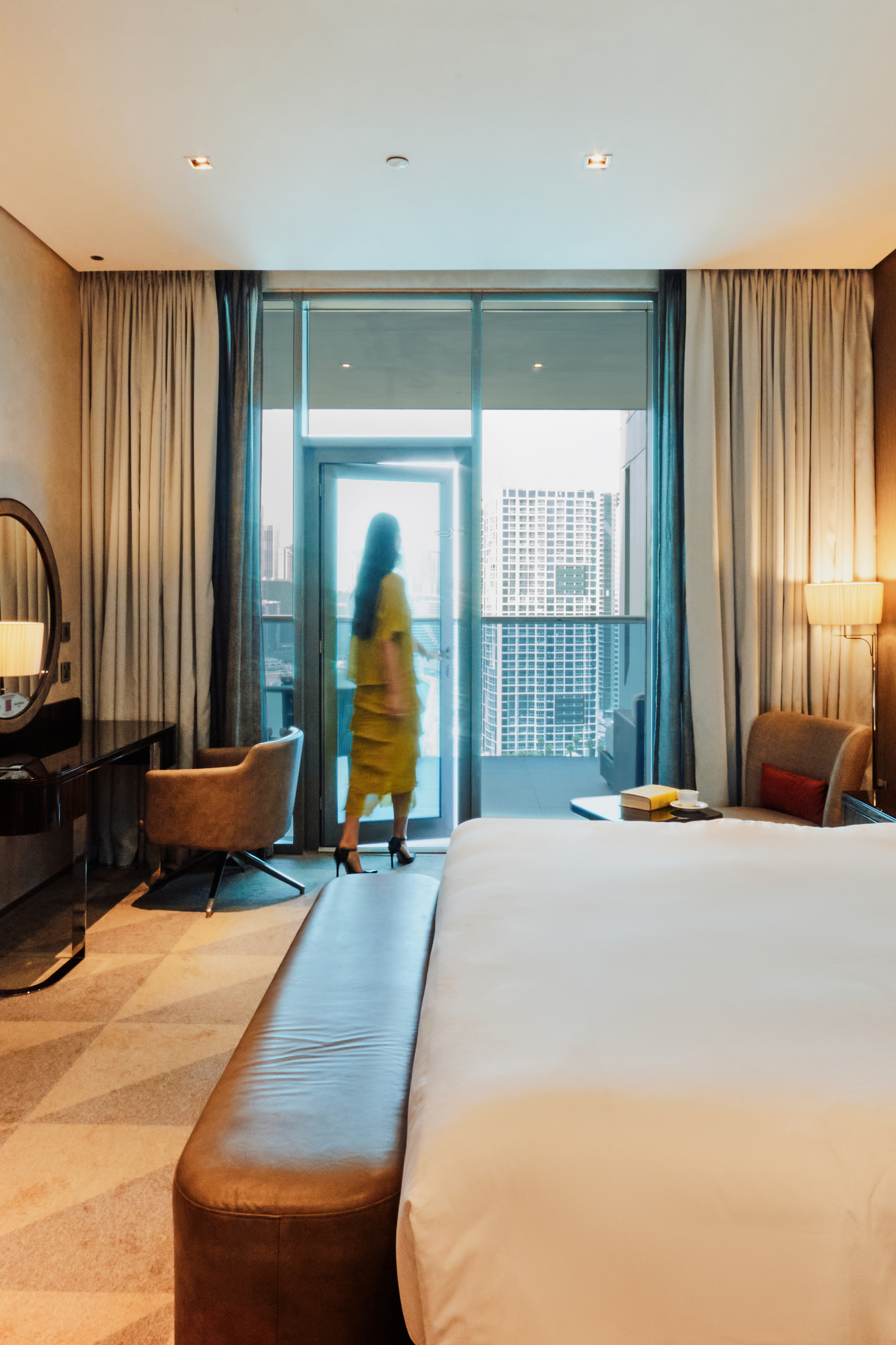 A woman in a hotel room, stepping towards the balcony door with one hand on the handle, looking out at the view beyond.