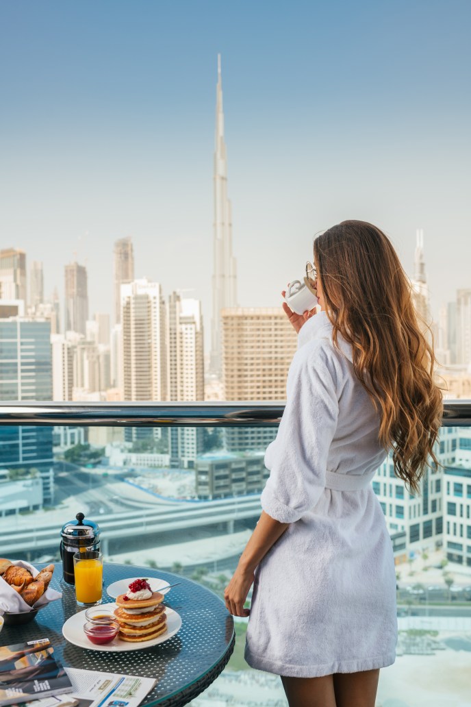 A woman in a bathrobe standing on a balcony, drinking from a cup and looking at the view, with a table set for breakfast next to her