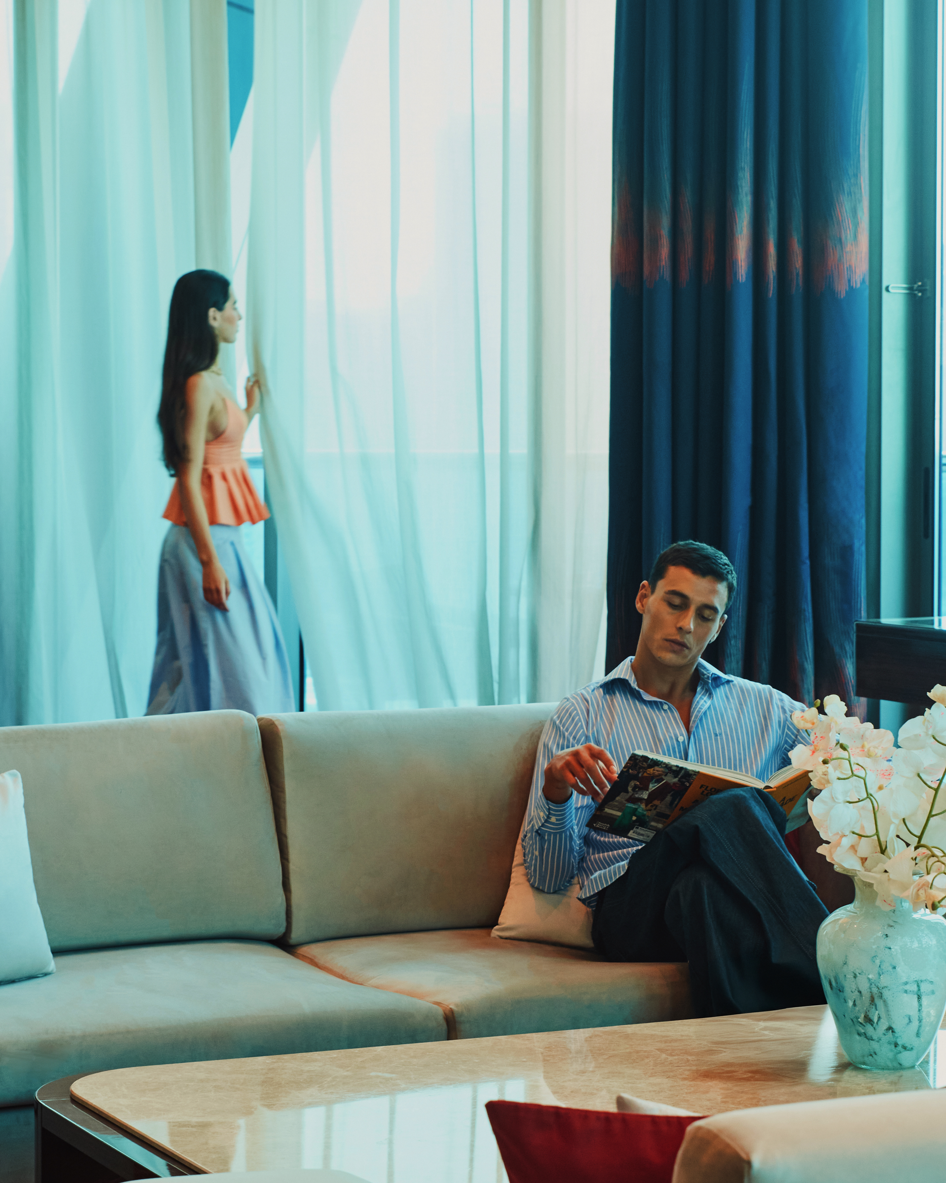 A man sitting on a couch, reading a book, while a woman stands next to the window.