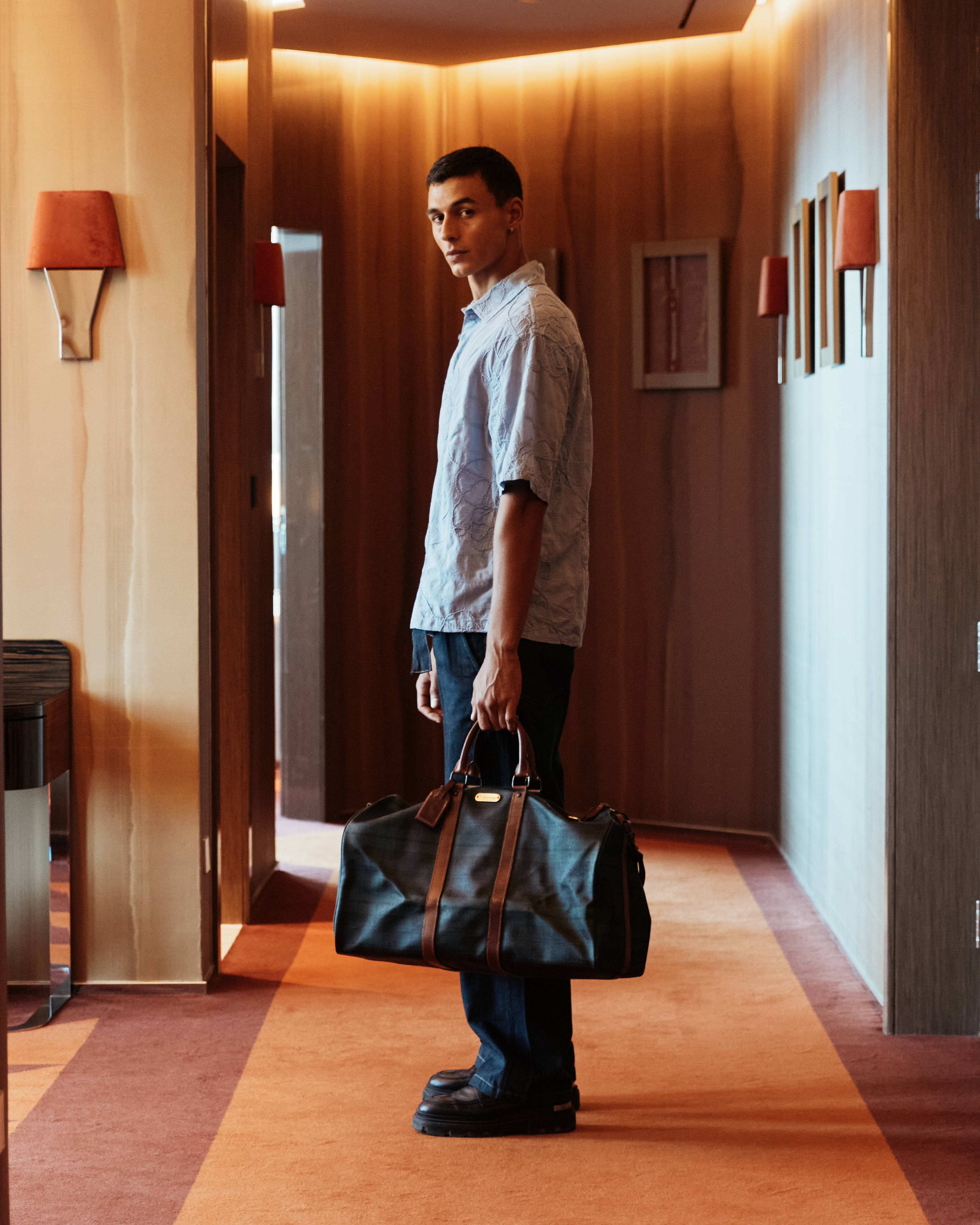 A man standing at the entrance of a hotel room, holding a travel bag.