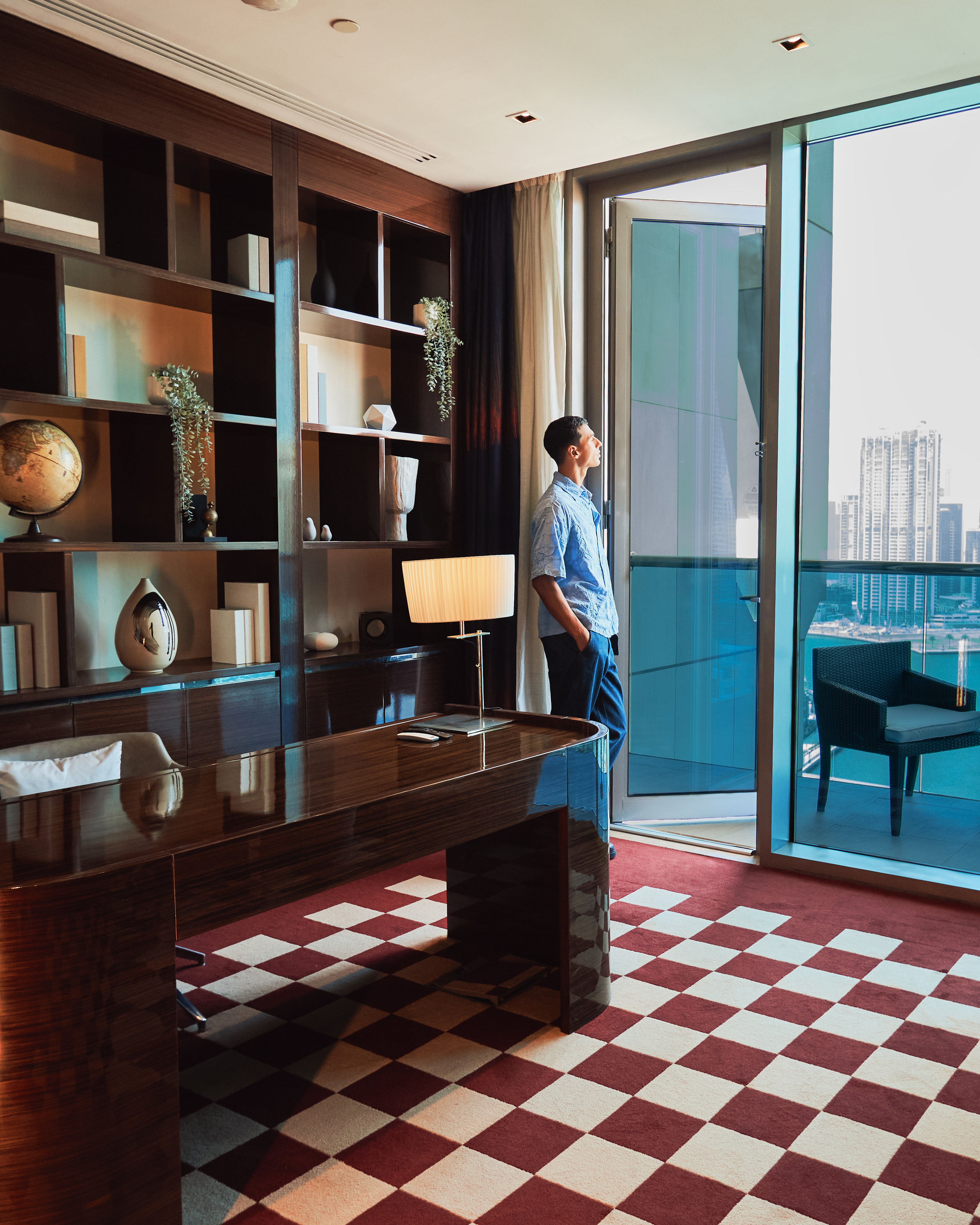 A man standing in the office/study of a penthouse, next to an open balcony door, looking at the city view.