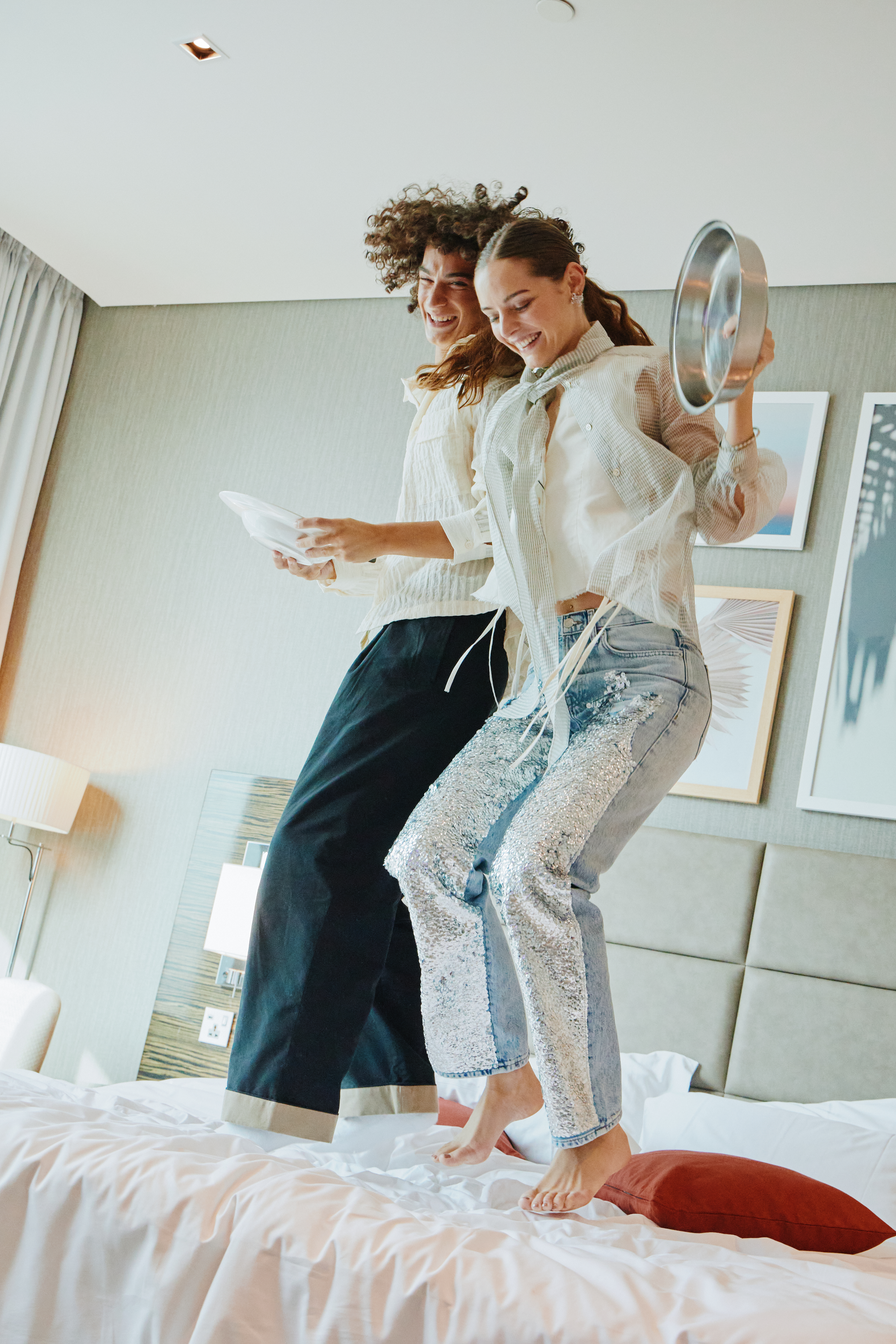 A youthful, happy couple jumping on a hotel bed, holding plates in their hands.